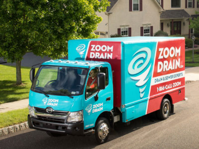A red and blue ZOOM DRAIN truck drives down asunny tree-lined street in a suburban neighborhood.