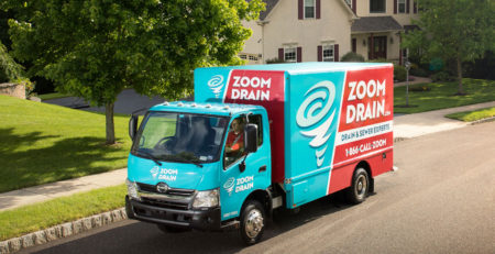 A red and blue ZOOM DRAIN truck drives down asunny tree-lined street in a suburban neighborhood.