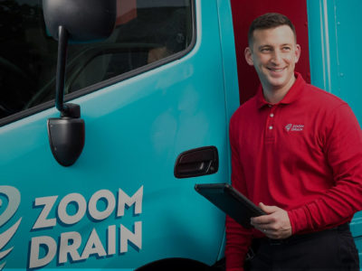 Man in a long-sleeved red ZOOM DRAIN shirt holding a tablet leans against a red and blue ZOOM DRAIN truck.
