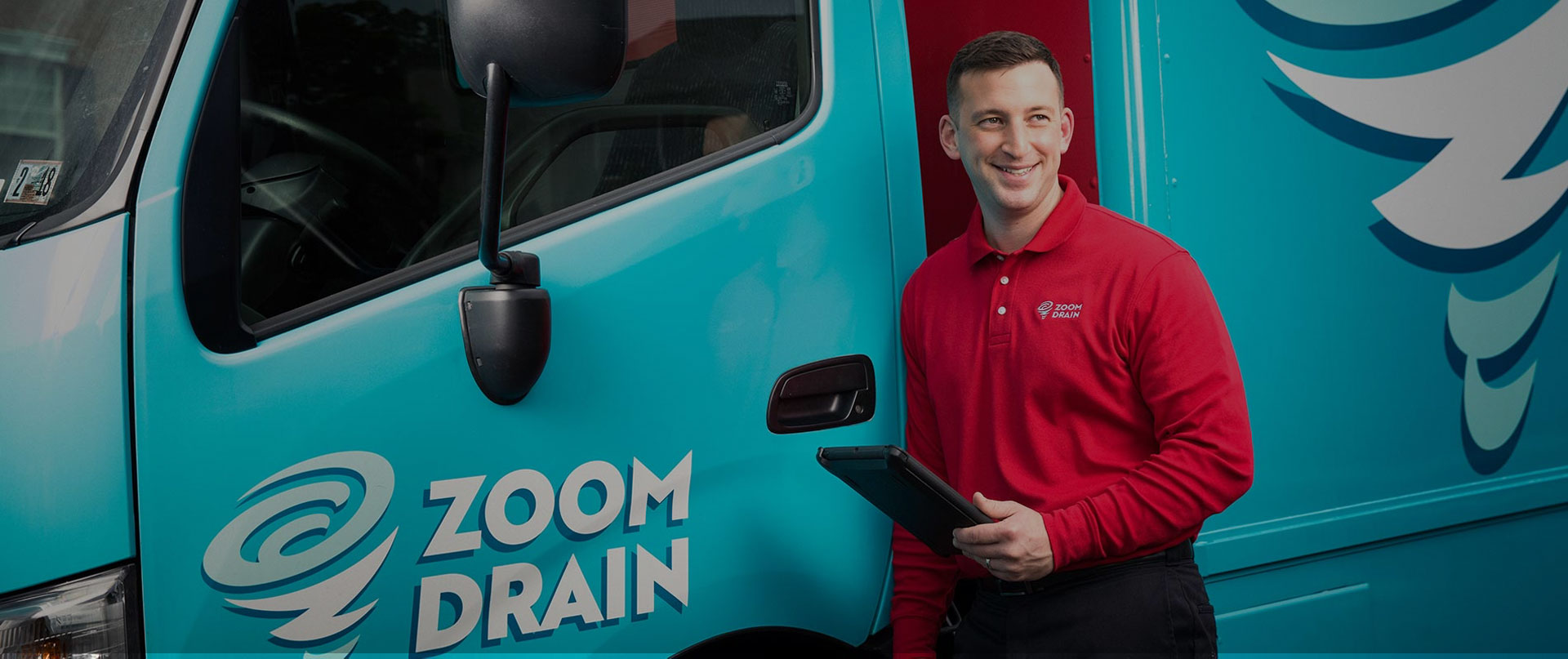Man in a long-sleeved red ZOOM DRAIN shirt holding a tablet leans against a red and blue ZOOM DRAIN truck.
