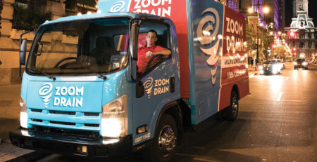 Smiling worker behind the wheel of a red and blue ZOOM DRAIN truck on a busy street at night